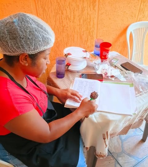 Voluntária de touca e avental anota informações em um caderno, sentada a uma mesa com marmitas e copos.